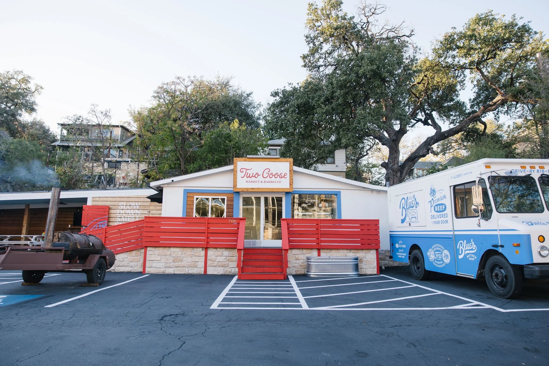 The Two Goose Market & Barbecue storefront at 706 N Lamar in Austin, with the red fence, smoker trailer, and live oak canopy