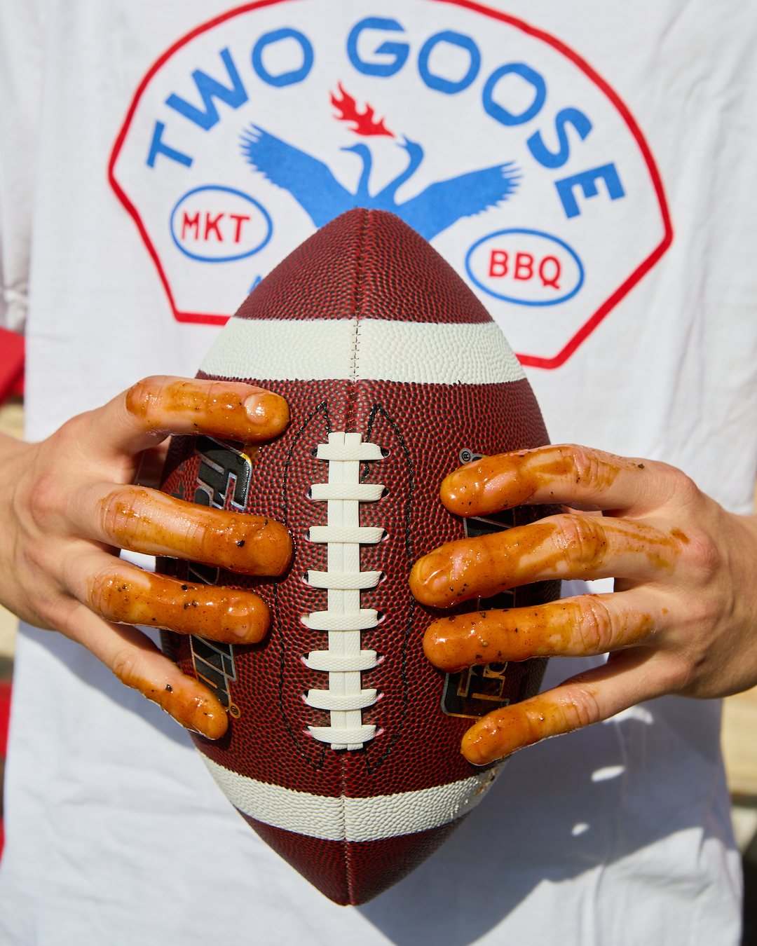 Saucy hands gripping a football in front of a Two Goose Market & Barbecue shirt